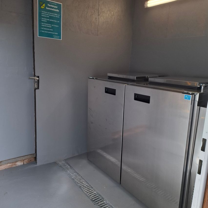 Ein leerer Raum mit Edelstahlkühlschränken, einer Tür und einem Wandschild in Deutsch.
