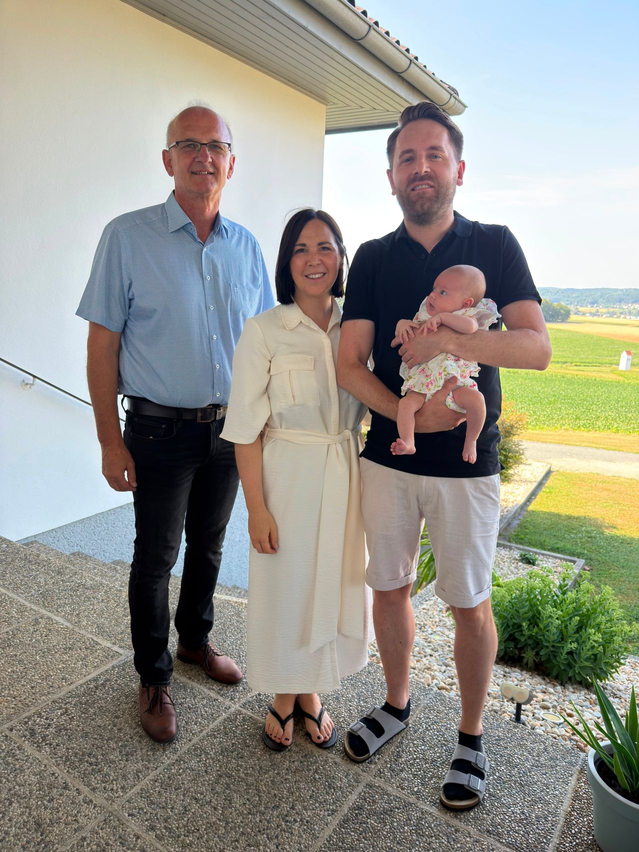 Drei Personen stehen auf einer Veranda mit einem Baby. Der Mann links trägt ein blaues Hemd, die Frau in der Mitte trägt ein weißes Kleid und der Mann rechts hält ein Baby.