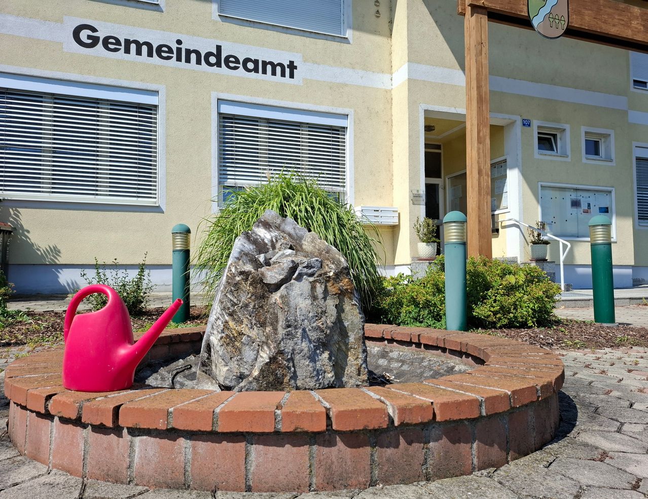 Ein großer Stein in einer runden Ziegelpflanzung mit einer rosa Gießkanne vor einem Gebäude, das mit 'Gemeindeamt' beschriftet ist.