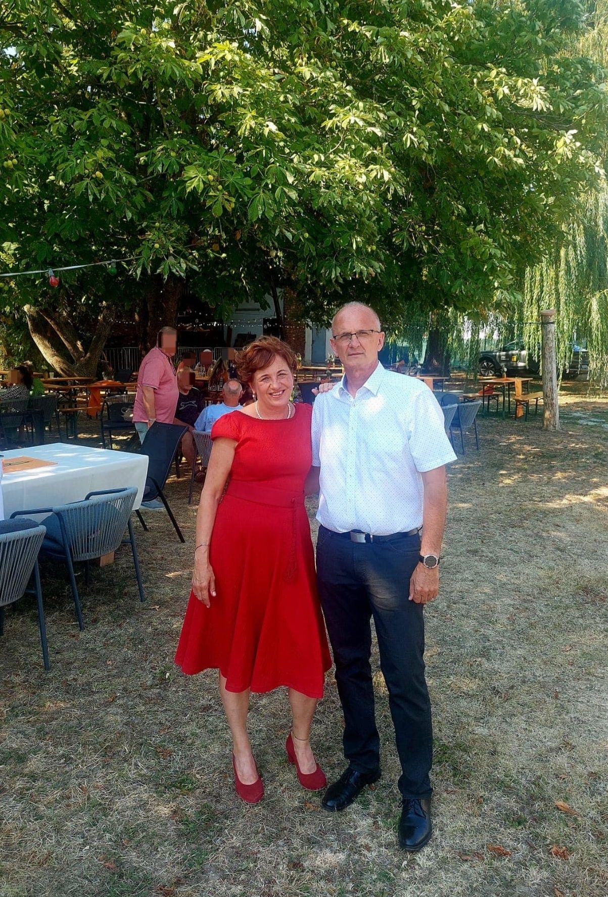 Ein Mann und eine Frau stehen lächelnd für ein Foto in einem Outdoor-Essbereich. Die Frau trägt ein rotes Kleid, und der Mann trägt ein weißes Hemd und dunkle Hosen. Hinter ihnen sind Tische und Stühle unter Bäumen aufgestellt.