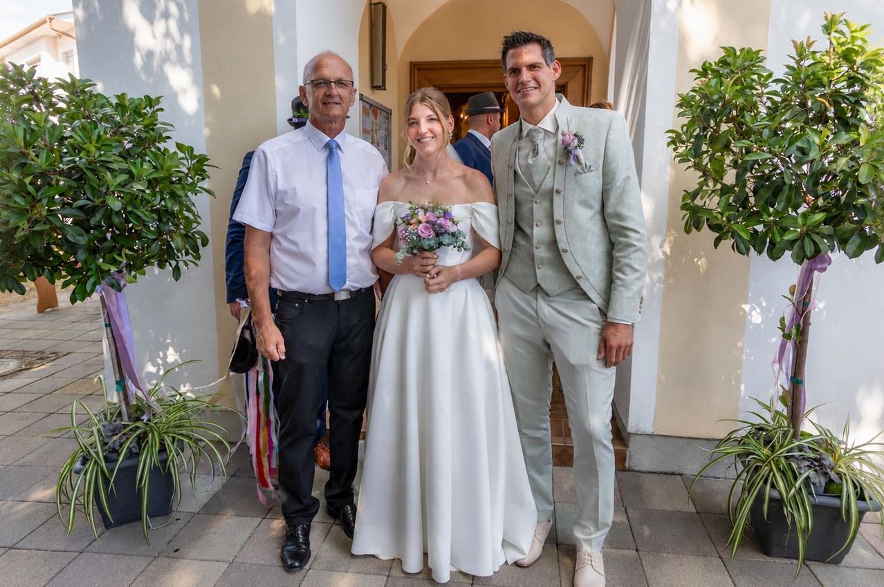 Eine Braut in einem weißen Kleid und ein Bräutigam in einem hellgrauen Anzug stehen mit einem Mann in einer blauen Krawatte, alle lächeln vor einem Gebäude.