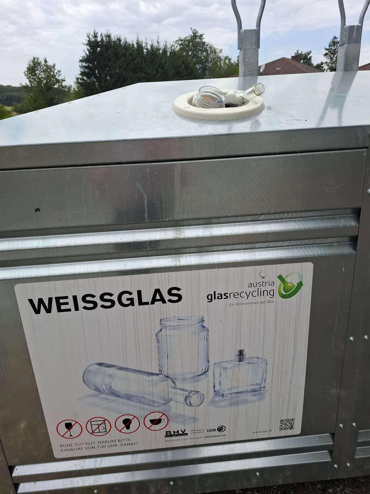 Ein Metallrecyclingbehälter mit einem Schild, das 'Weissglas' und eine Illustration von Glasflaschen und Gläsern zeigt. Das Schild ist auf Deutsch und zeigt ein grünes Recyclingsymbol.