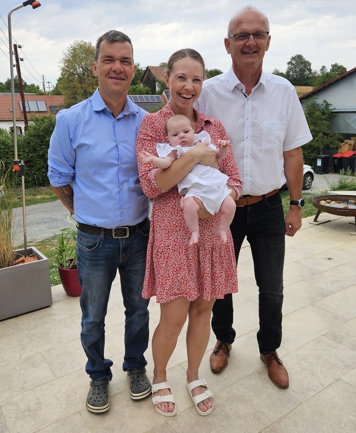 Drei Menschen stehen lächelnd zusammen auf einer Terrasse. Eine Frau hält ein Baby, und zwei Männer stehen auf beiden Seiten. Dahinter stehen Pflanzen, ein Feuerkorb und ein Haus.