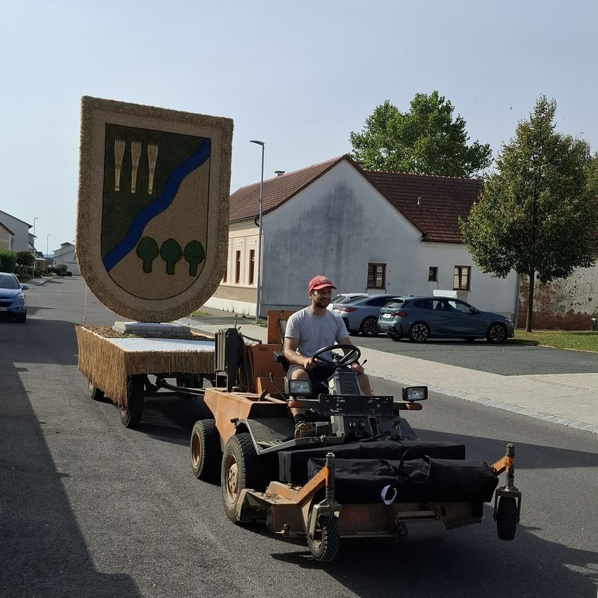 Ein Mann fährt einen großen Traktor mit einem Anhänger, der ein Wappen trägt, eine Straße hinunter, gesäumt von geparkten Autos und Gebäuden.