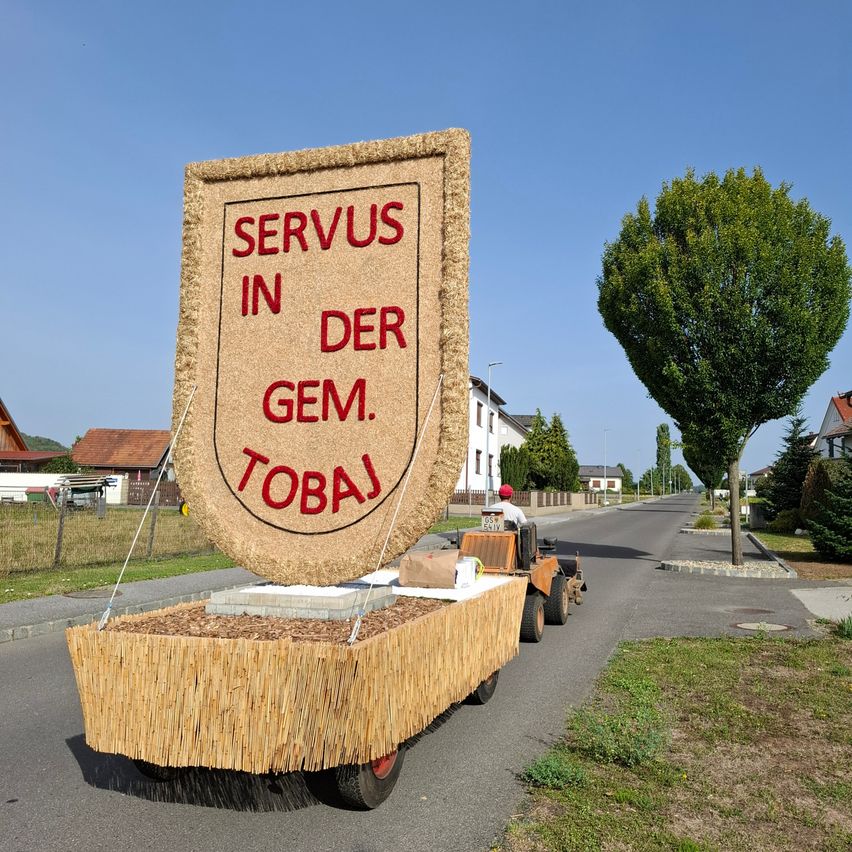 Ein großer Heuballen in Form eines Schildes mit rotem Text, der 'SERVUS IN DER GEM. TOBAJ' lautet, wird von einem Traktor auf einer Straße gezogen.