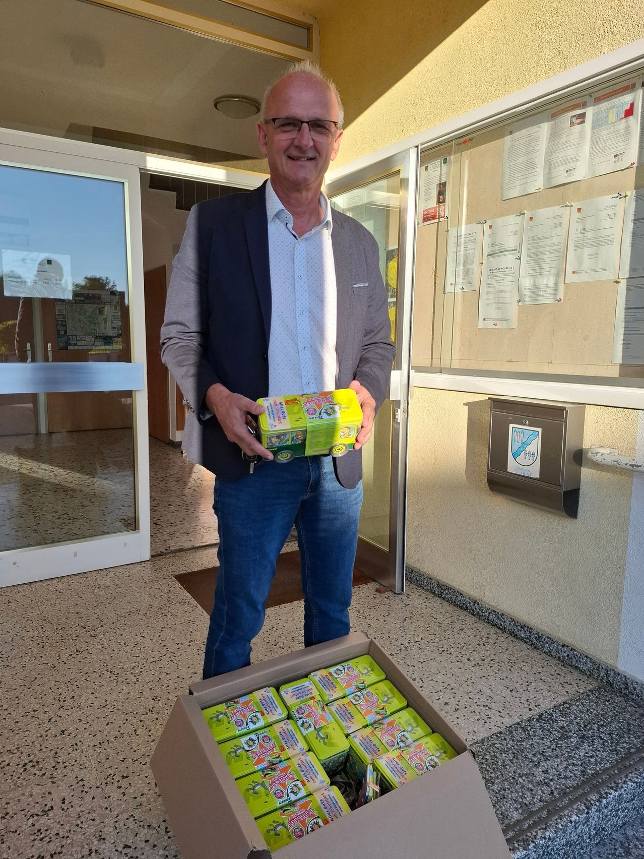 Ein Mann in einem grauen Blazer und blauen Jeans hält eine Schachtel mit gelben und grünen Gegenständen, lächelt und steht vor einer Glastür.