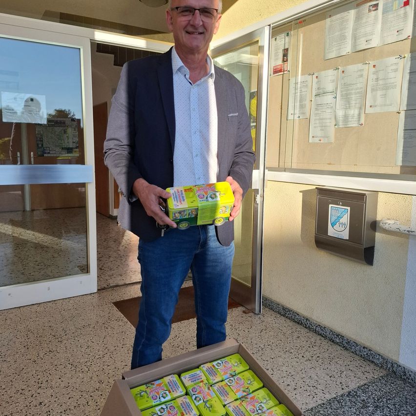 Ein Mann in einem grauen Blazer und blauen Jeans hält eine Schachtel mit gelben und grünen Gegenständen, lächelt und steht vor einer Glastür.