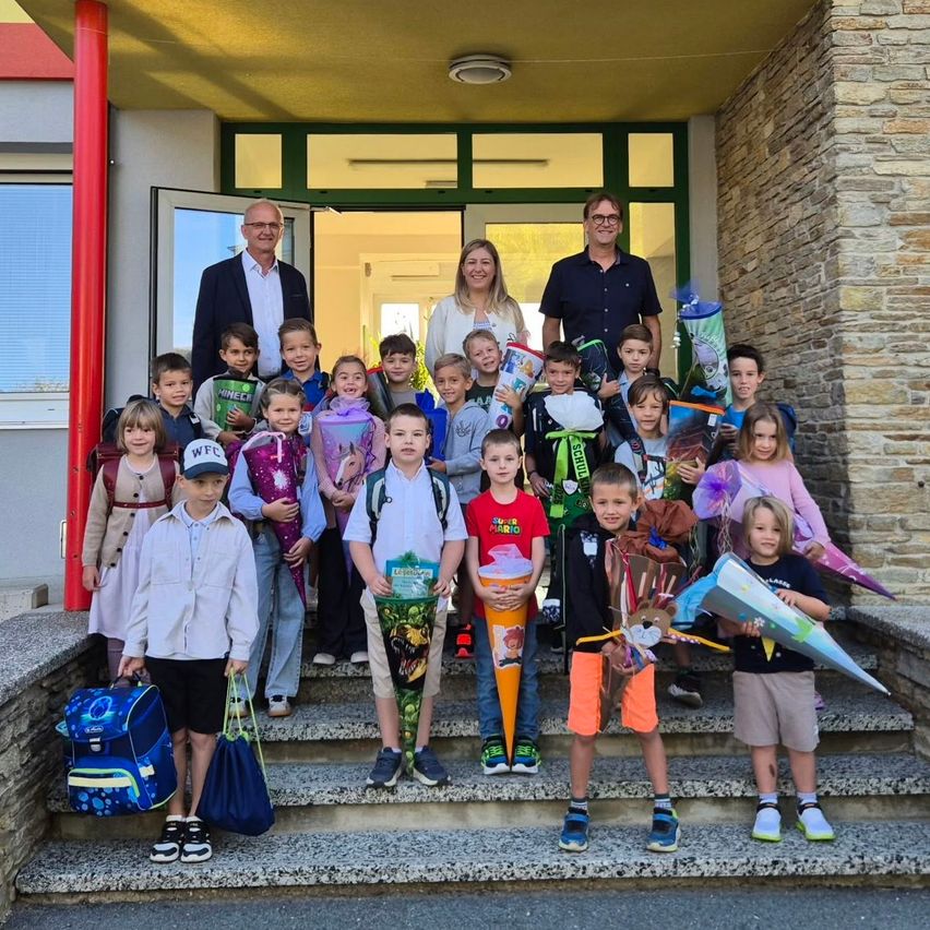 Eine Gruppe von Kindern und zwei Erwachsenen steht auf den Stufen eines Gebäudes, hält verschiedene Geschenke und trägt Schuluniformen. Die Erwachsenen lächeln und scheinen Lehrer zu sein.