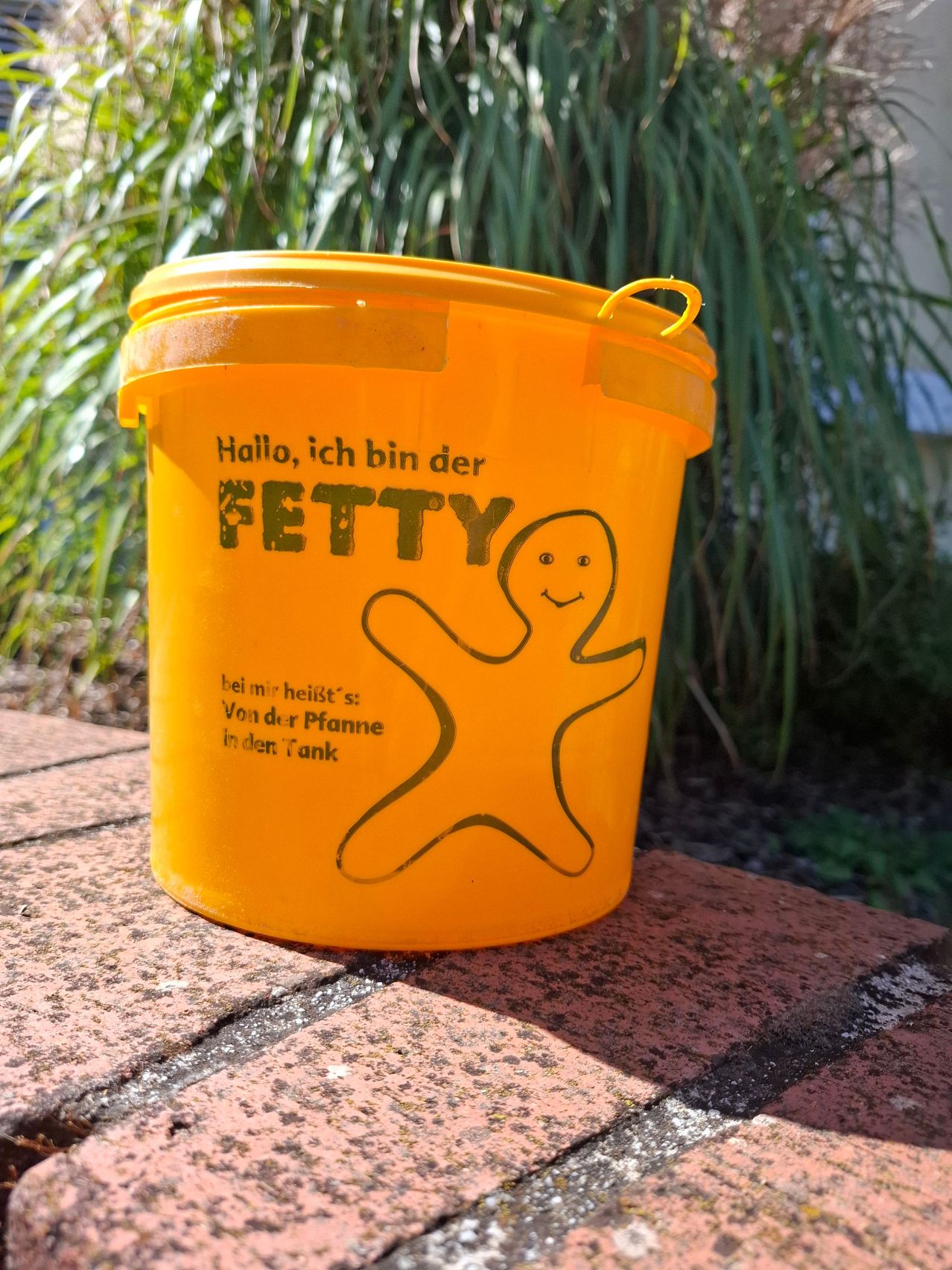 Ein orangefarbener Eimer mit den Worten 'Hallo, ich bin der FETTY' und einer Cartoon-Figur darauf. Der Eimer steht auf einer Ziegelfläche.