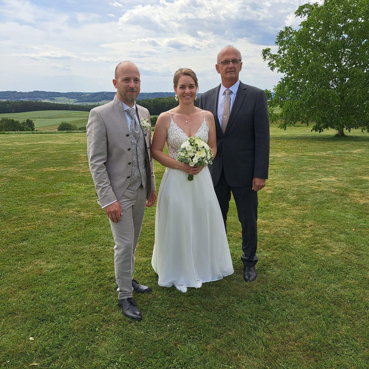 A bride in a white gown and two groomsmen in suits stand in a field with a bouquet and a tree in the background.