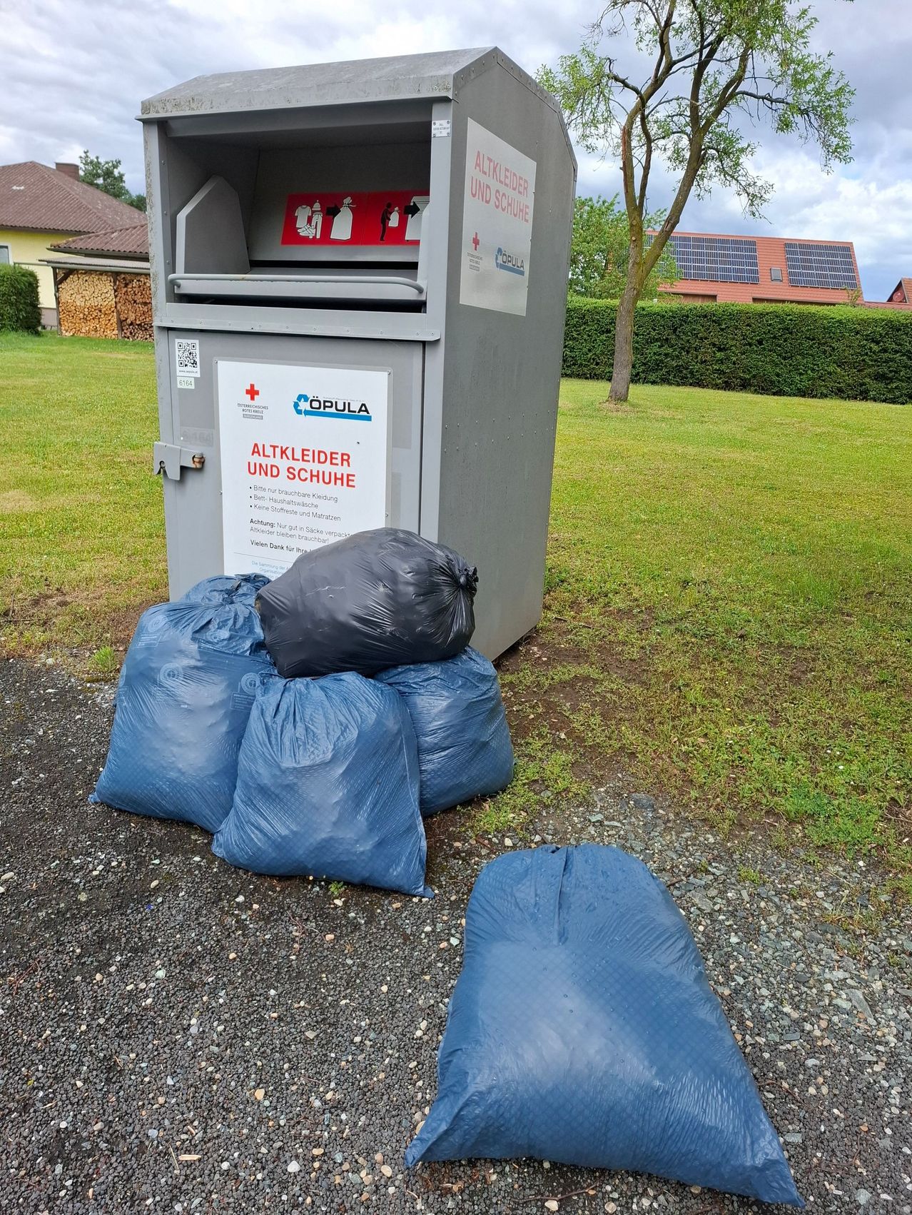 A donation box for old clothes and shoes with blue bags on the ground in front of it. Behind the box is a hedge, a tree, and a building with solar panels.