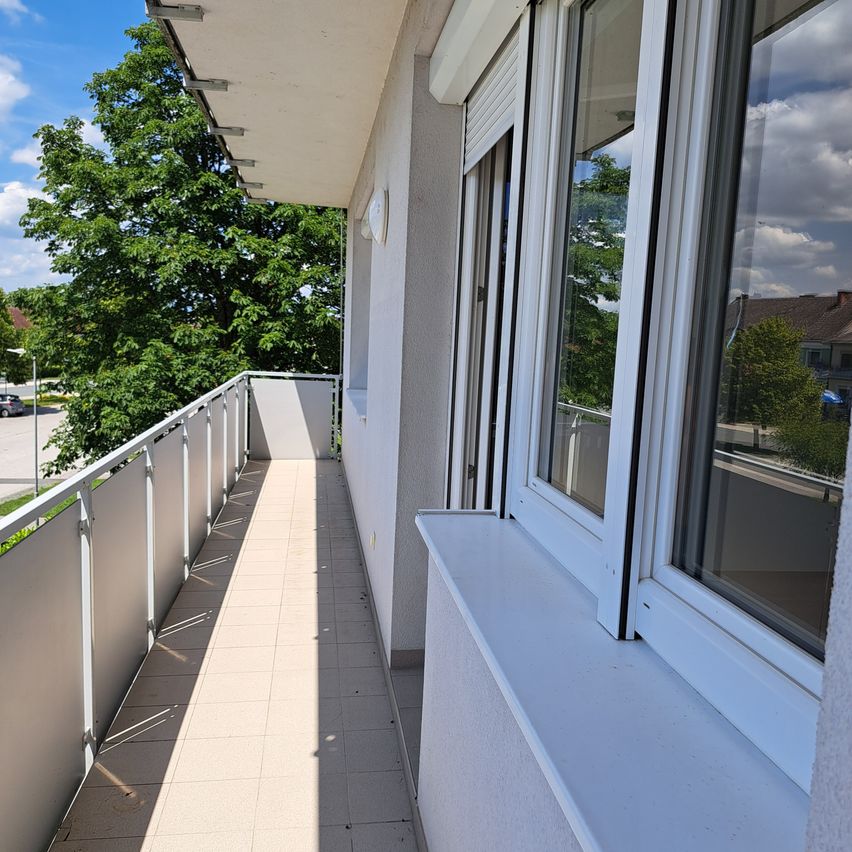 Ein Balkon mit gefliestem Boden und einem Metallgeländer. Es gibt zwei Fenster mit weißen Rahmen und einem weißen Fensterbrett. Der Balkon blickt auf einen Parkplatz mit Autos und Bäumen.
