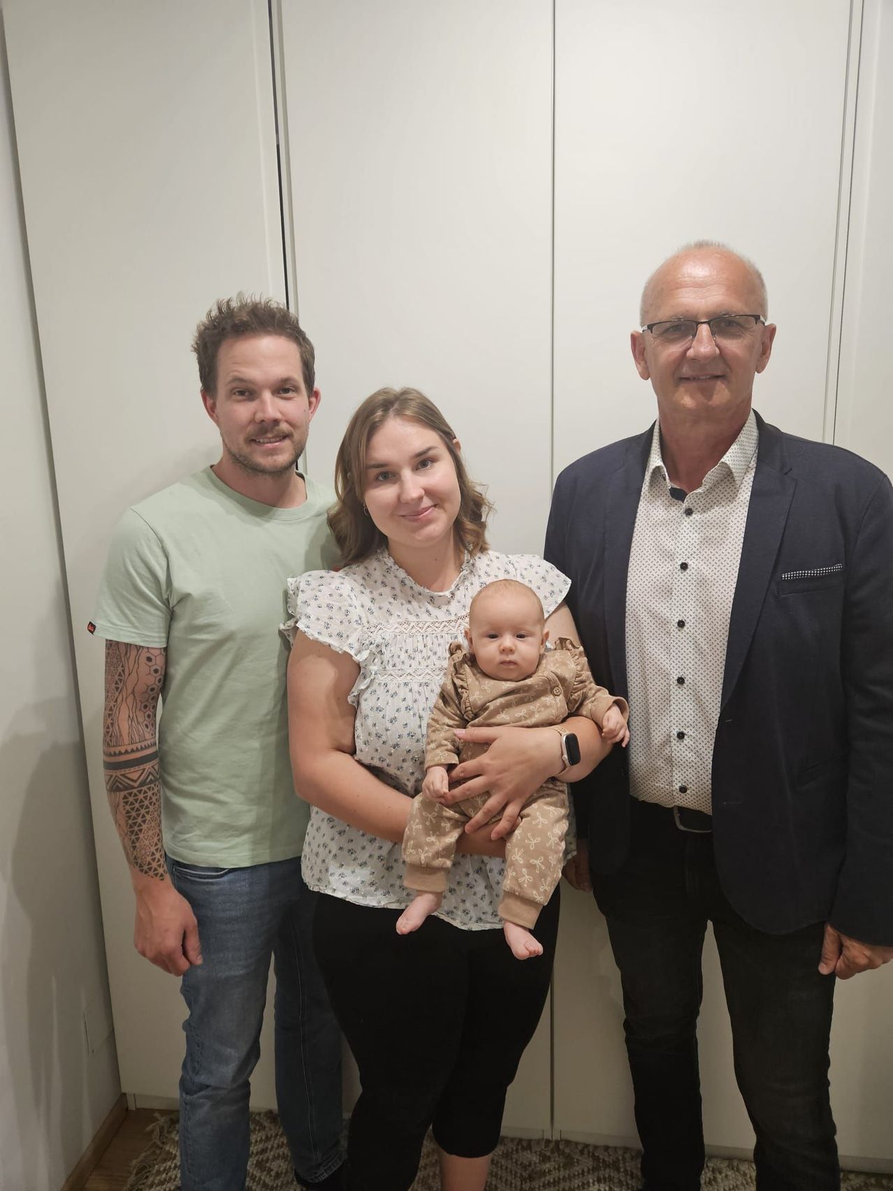 Drei Menschen stehen eng beieinander und lächeln. Die Frau hält ein Baby, während die beiden Männer zu beiden Seiten stehen. Sie scheinen für ein Foto zu posieren.