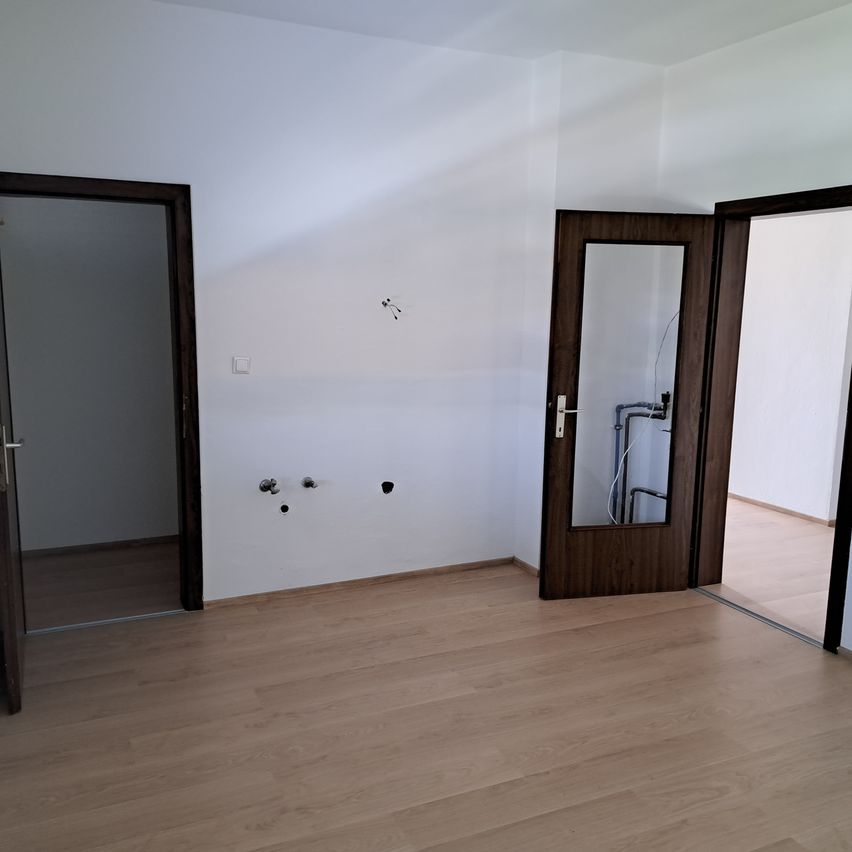 Ein leerer Raum mit Holzboden und weißen Wänden. Es gibt drei Türen, von denen eine offen ist. Die Wände haben einige sichtbare Schrauben und eine Steckdose.