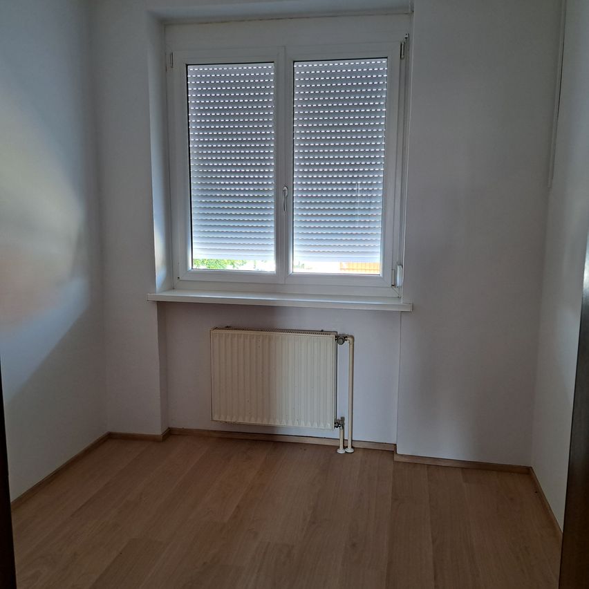 Ein leerer Raum mit weißen Wänden und Holzboden. Ein Fenster mit weißen Jalousien ist teilweise geöffnet und gewährt einen Blick nach draußen. Ein weißer Heizkörper ist unterhalb des Fensters an der Wand montiert.