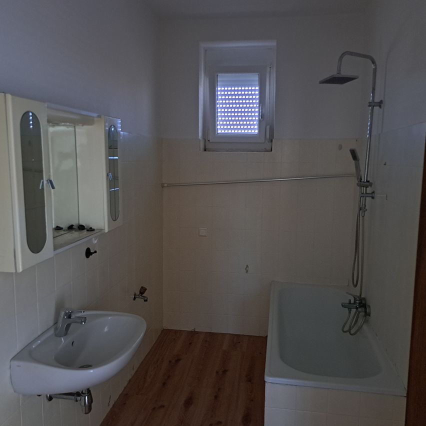 Ein leeres Badezimmer mit Badewanne, Waschbecken und Spiegel. Die Wände und der Boden sind gefliest. Ein Duschkopf ist an einem Metallarm befestigt. Es gibt ein Fenster mit Jalousien.