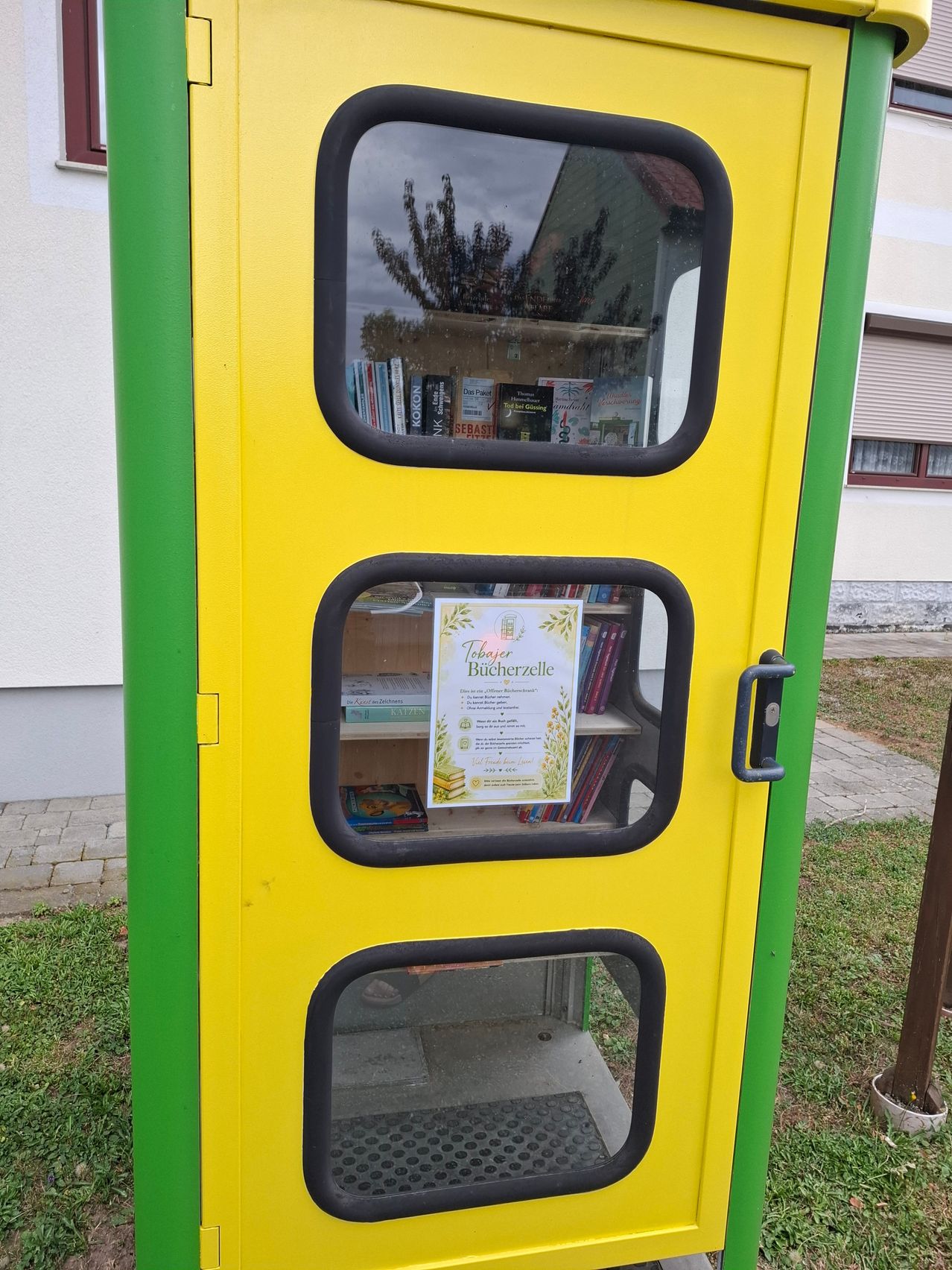 Ein gelber und grüner öffentlicher Bücherschrank mit drei Glastüren und einem Schild mit dem Text Taker Bucherzelle. Er hat Regale im Inneren mit Büchern. Er steht auf einem Rasenbereich.