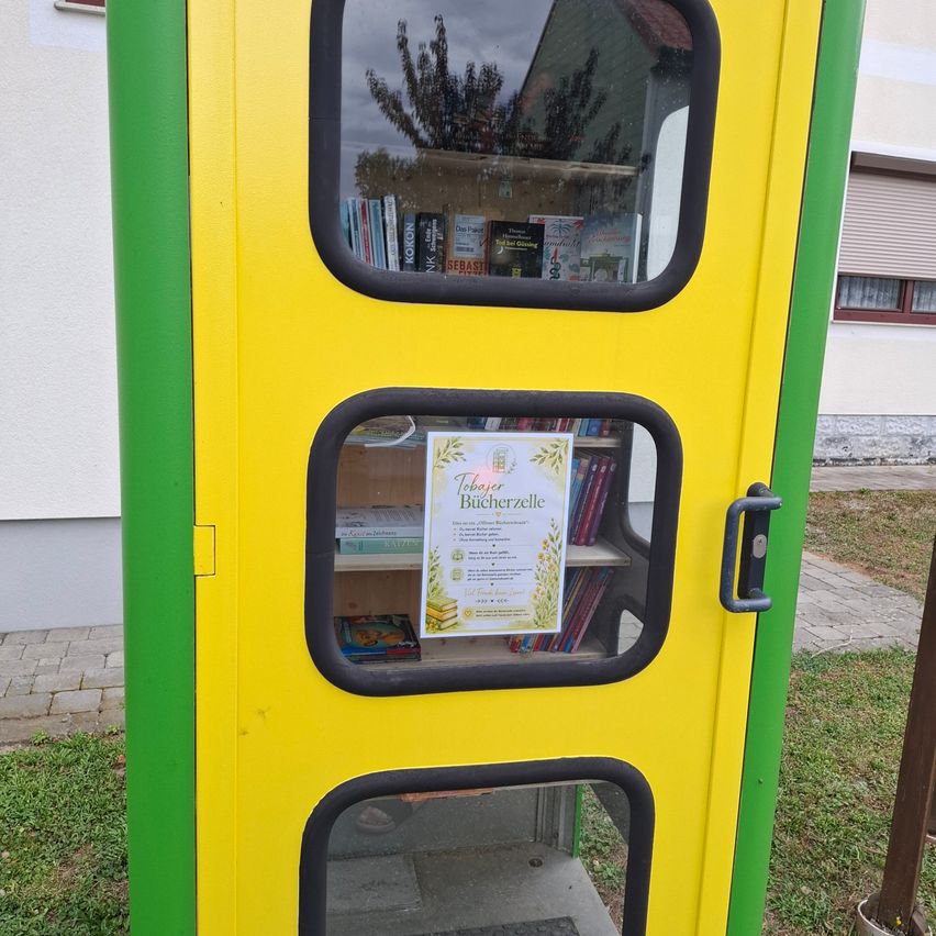 Ein gelber und grüner öffentlicher Bücherschrank mit drei Glastüren und einem Schild mit dem Text Taker Bucherzelle. Er hat Regale im Inneren mit Büchern. Er steht auf einem Rasenbereich.