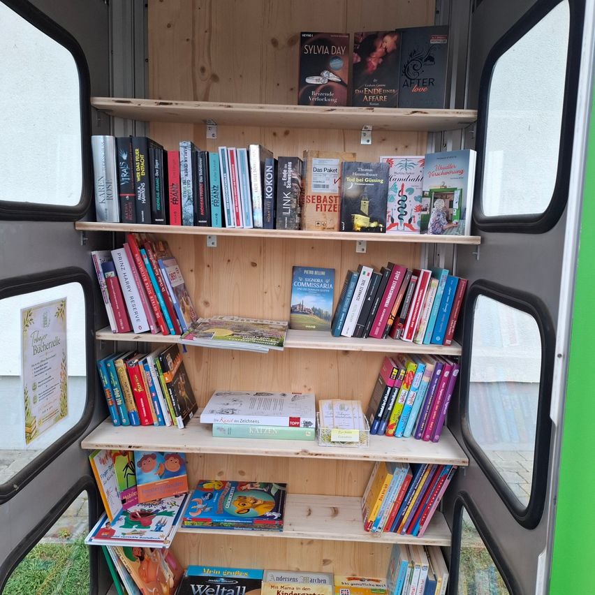 Ein Bücherregal mit verschiedenen Büchern, darunter Romane und Kinderbücher, befindet sich in einem Bus. Die Regale sind aus Holz und die Bücher sind ordentlich angeordnet. Der Bus hat Glasfenster auf beiden Seiten.