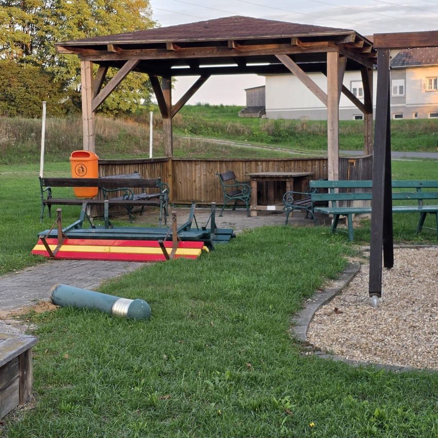 Ein hölzerner Pavillon mit Bänken und einem Tisch, einem Mülleimer und einem Weg, der zu einem Rasenbereich führt.