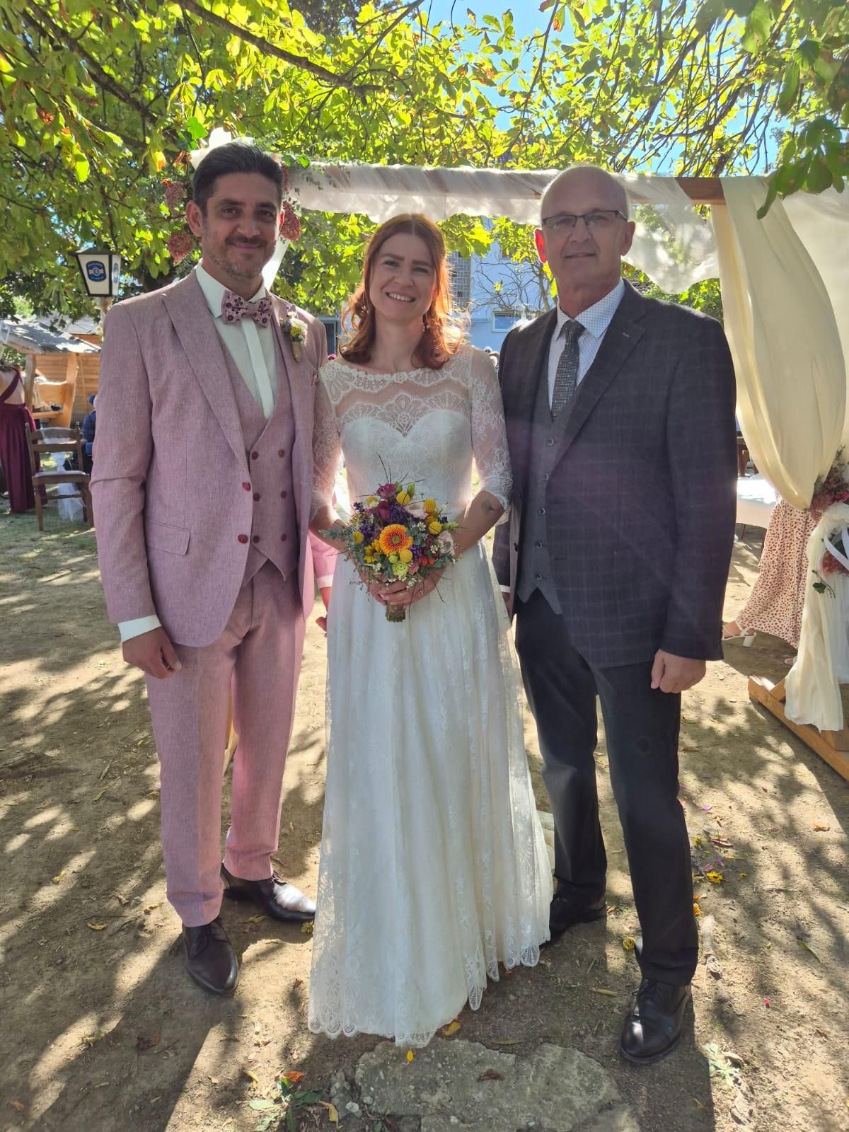 Ein frisch verheiratetes Paar und ein Mann stehen zusammen unter einem Baldachin und lächeln für ein Foto. Der Bräutigam trägt einen rosa Anzug, die Braut ein weißes Hochzeitskleid und der Mann einen grauen Anzug.
