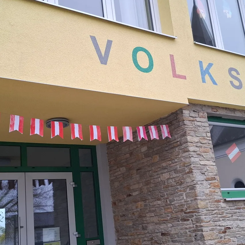 Die Vorderseite eines Gebäudes mit dem Wort Volksschule in bunten Buchstaben. Kleine Flaggen mit roten und weißen Streifen hängen am Dach.