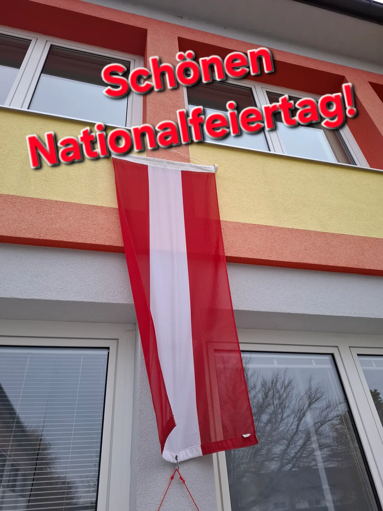 Eine rot-weiße Fahne hängt an einem Gebäude mit Balkon. Die Fahne ist mit einem roten Seil am Gebäude befestigt. Das Gebäude hat ein Glasfenster mit Jalousien und eine gelbe Wand. Der Text an der Wand lautet 'Schönen Nationalfeiertag!'