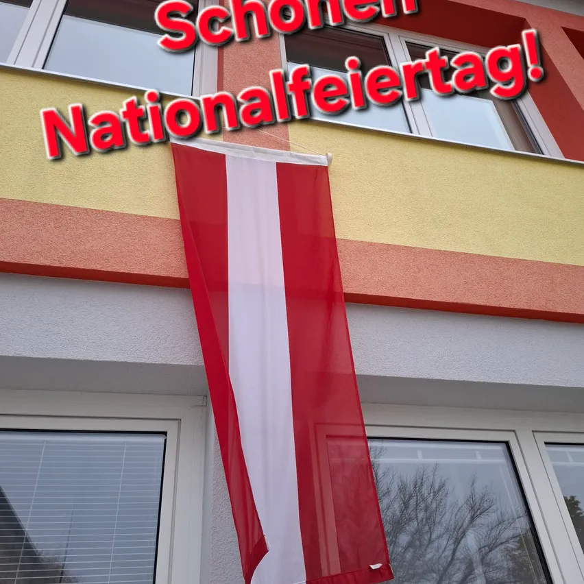 Eine rot-weiße Fahne hängt an einem Gebäude mit Balkon. Die Fahne ist mit einem roten Seil am Gebäude befestigt. Das Gebäude hat ein Glasfenster mit Jalousien und eine gelbe Wand. Der Text an der Wand lautet 'Schönen Nationalfeiertag!'