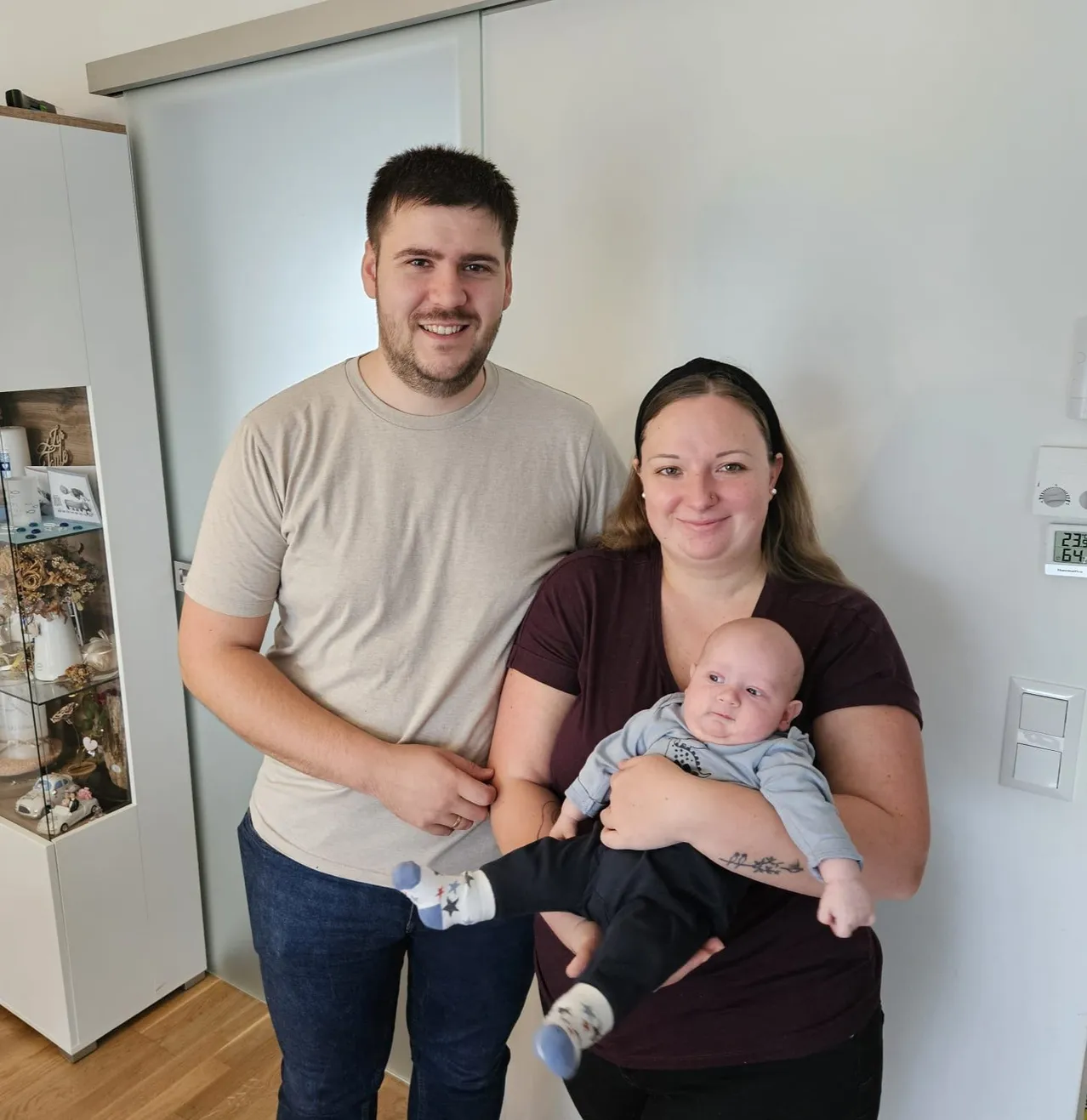 Ein Mann und eine Frau stehen in einem Raum mit einem Baby. Der Mann lächelt und hat seine Hand auf der Schulter der Frau. Die Frau hält ein Baby in ihren Armen und lächelt. Hinter ihnen ist eine weiße Wand mit einer Glastür und einem Schrank mit ausgestellten Gegenständen.