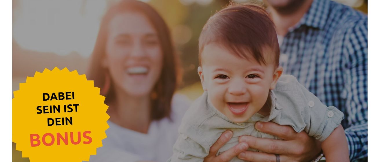 Ein lächelnder Vater hält ein Baby in seinen Armen, mit einer lächelnden Mutter im Hintergrund. Das Baby trägt ein graues Shirt und hat kurze braune Haare.