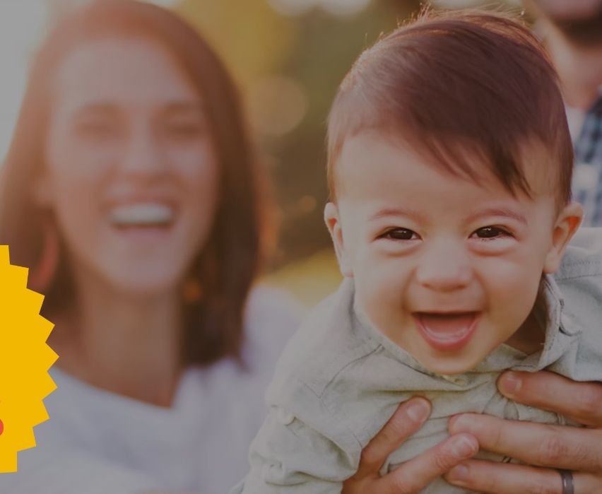 Ein lächelnder Vater hält ein Baby in seinen Armen, mit einer lächelnden Mutter im Hintergrund. Das Baby trägt ein graues Shirt und hat kurze braune Haare.