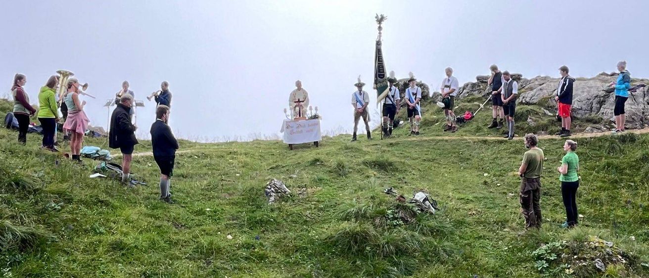 Eine Gruppe von Menschen versammelt sich auf einem grasbewachsenen Hügel mit einer Fahne. Ein Tisch ist in der Nähe der Felsen aufgestellt, mit einigen Personen, die in der Nähe stehen. Der Himmel ist bedeckt.