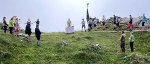 Eine Gruppe von Menschen versammelt sich auf einem grasbewachsenen Hügel mit einer Fahne. Ein Tisch ist in der Nähe der Felsen aufgestellt, mit einigen Personen, die in der Nähe stehen. Der Himmel ist bedeckt.