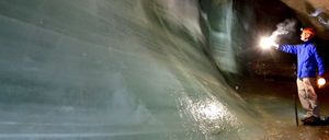 Ein Mann in einer blauen Jacke und Helm steht in einer Höhle mit einer leuchtenden Taschenlampe, die die eisigen Wände und den Boden beleuchtet.