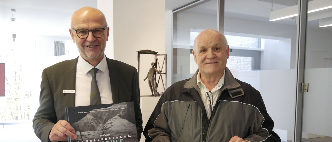Zwei Männer halten ein Buch und ein Schild. Das Buch heißt Schellenberger Eishohle. Das Schild sagt Gemeinsam ist einfach. 2.000,- €. Dahinter ist eine Glaswand und eine Skulptur.