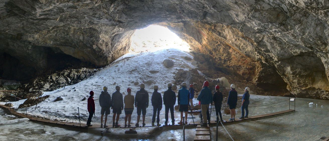 Eine Gruppe von Menschen in Helmen und Jacken steht in einer Höhle und schaut zum Licht am Eingang. Holzstege verbinden sie mit dem Höhlenboden.