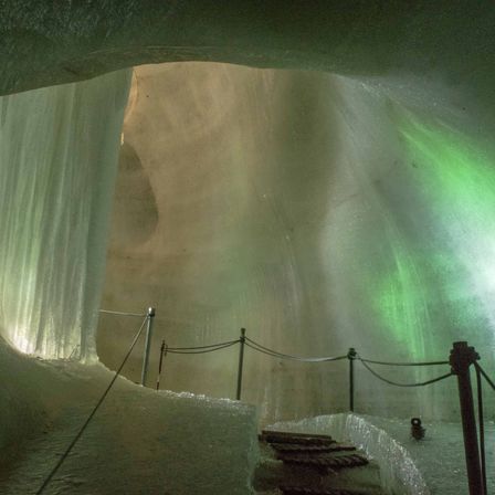 Ein Blick in eine Eishöhle mit einem leuchtend grünen Licht auf der rechten Seite. Stufen führen zu einer tieferen Ebene, und Geländer sind für die Sicherheit vorhanden.