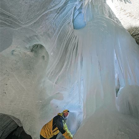Ein Mann in einem gelben und schwarzen Jackett klettert an einer Eiswand in einer Höhle hoch.