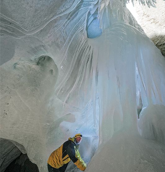 Ein Mann in einem gelben und schwarzen Jackett klettert an einer Eiswand in einer Höhle hoch.