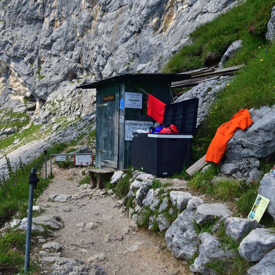 Eine kleine Hütte mit einer roten Flagge, die draußen hängt, in der Nähe eines felsigen Bergpfades. Die Hütte hat einen offenen Behälter, und es liegen verstreute Kleidungsstücke auf den Felsen.