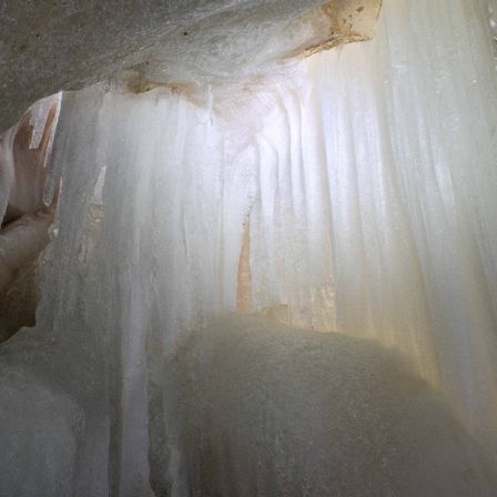 Eine Nahaufnahme einer eisigen Höhle mit Eiszapfen, die von der Decke hängen und von einer Lichtquelle beleuchtet werden.