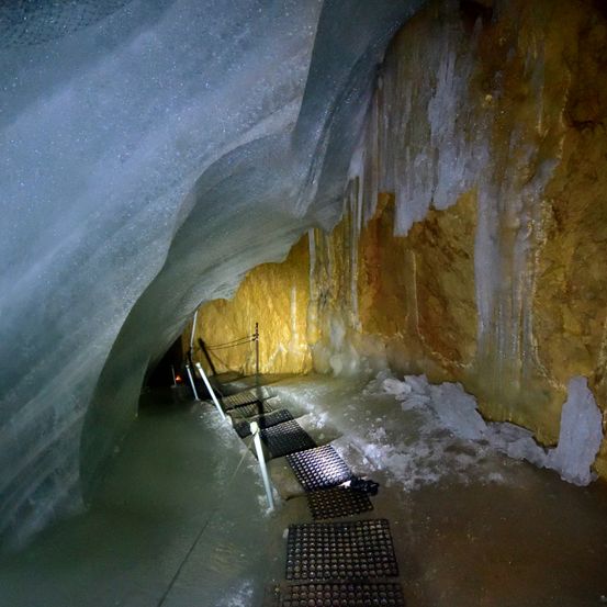 Ein eisiger Tunnel mit einem Gehweg aus Metallgittern. Die Wände bestehen aus einer Mischung aus Eis und Stein, mit Eiszapfen, die von der Decke hängen.