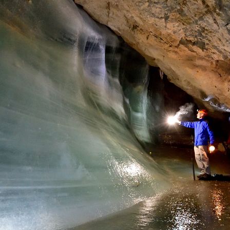 Ein Mann in einer blauen Jacke und einem orangefarbenen Helm erkundet eine große Eishöhle. Er hält eine Taschenlampe in der Hand und beleuchtet die eisigen Wände.