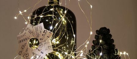 A black bottle adorned with golden lights and a small paper music note sits next to a small crocheted Christmas tree, both glowing with festive light strings.