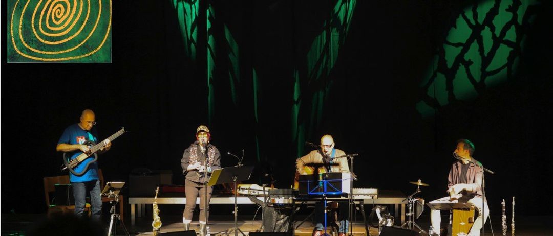 Eine Band tritt mit einem grünen Hintergrund auf der Bühne auf, bestehend aus einem Gitarristen, einem Keyboarder, einem Klarinettisten und einem Perkussionisten. Sie werden von grünen Bühnenlichtern beleuchtet.