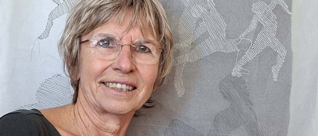 An older woman with glasses and a smile poses in front of a wall with an abstract drawing of people running.