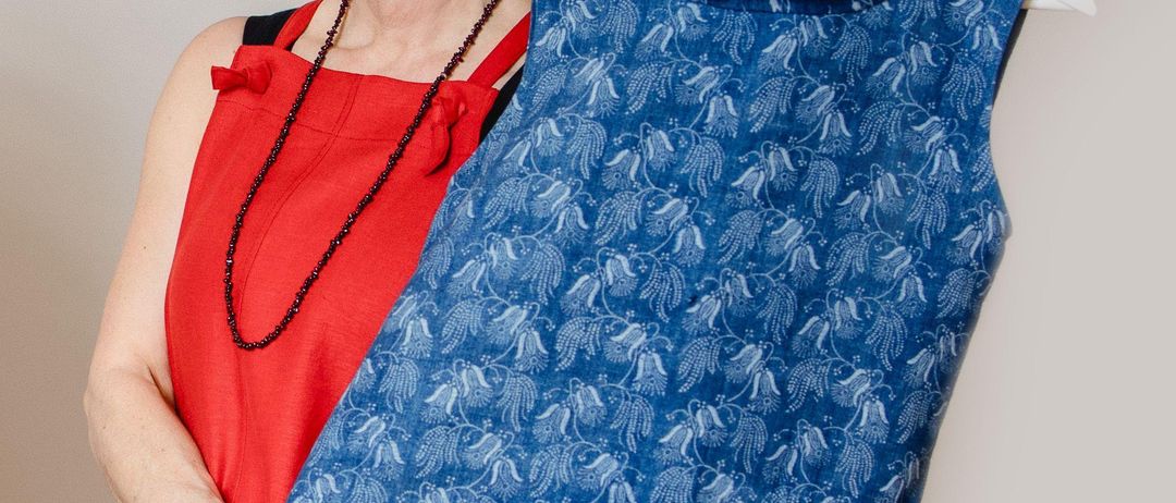 A woman in a red dress is holding a blue patterned sleeveless top on a hanger. She smiles and poses for a photo.