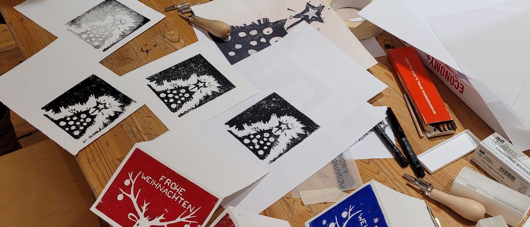 Ein Holztisch mit verschiedenen Kunstmaterialien und gedruckten Karten. Eine Person arbeitet an einer roten Karte mit einem Hirschdesign.