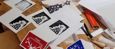 Ein Holztisch mit verschiedenen Kunstmaterialien und gedruckten Karten. Eine Person arbeitet an einer roten Karte mit einem Hirschdesign.