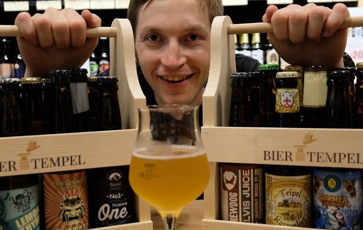 Ein lächelnder Mann hält zwei Holzkisten voller verschiedener Bierflaschen, eine mit einem Glas Bier davor. Die Kisten sind mit 'Bier Tempel' und 'Unsere Leidenschaft' beschriftet.